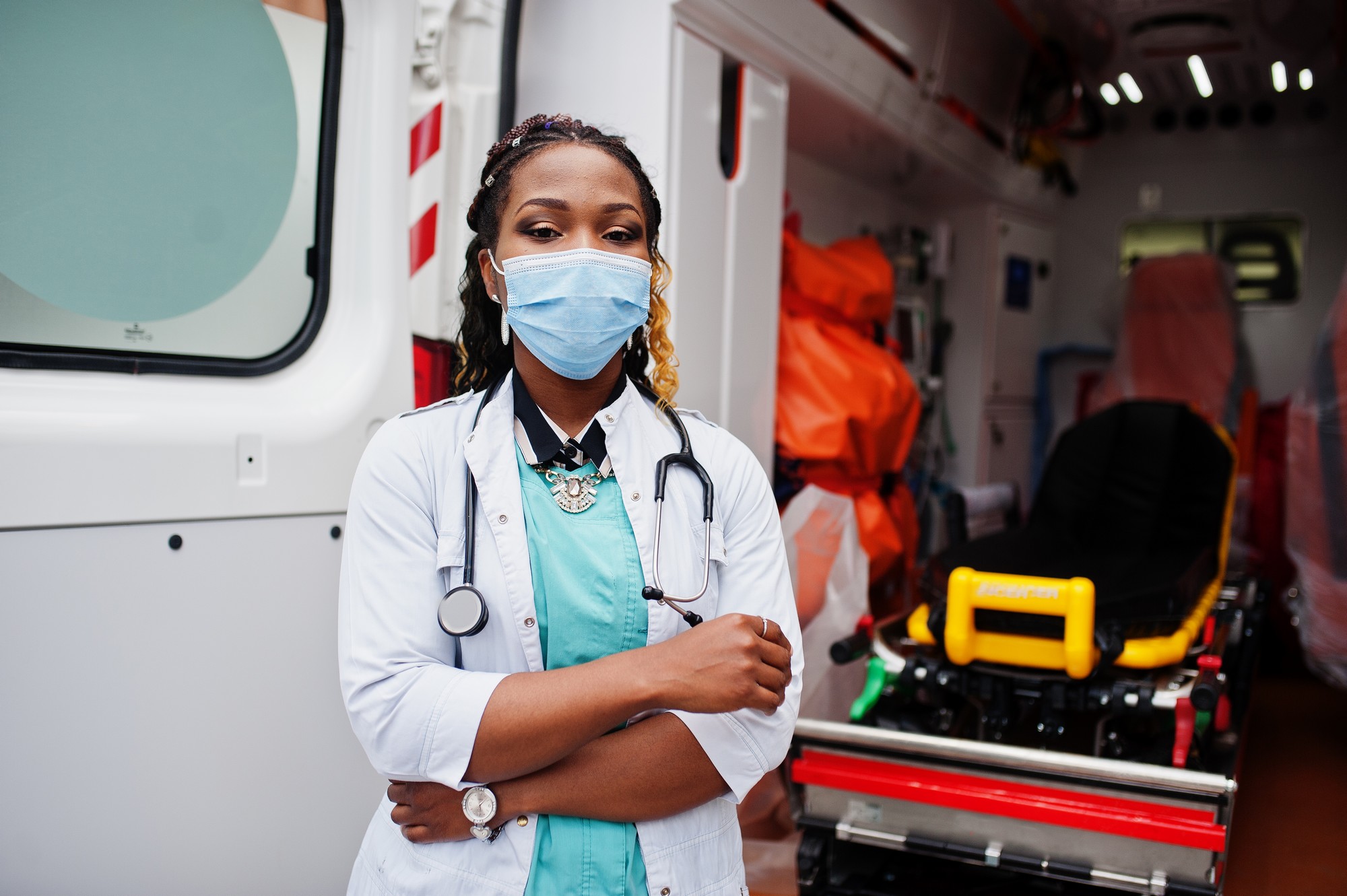 African american female paramedic in face protective medical mask standing in front of ambulance car.